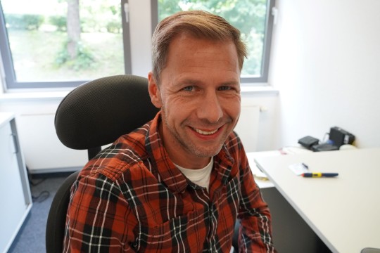 Ein Mann sitzt an seinem Arbeitsplatz und lächelt in die Kamera.