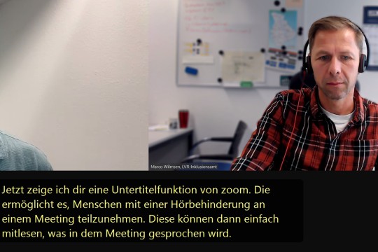 Ein Mann mit Headset sitzt am Computer und befindet sich in einem Zoom-Meeting. Die Untertitelfunktion von Zoom ist zu erkennen.