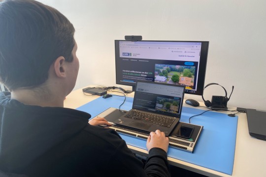 Eine Frau sitzt vor einem Laptop mit Braillezeile sowie einem Monitor.