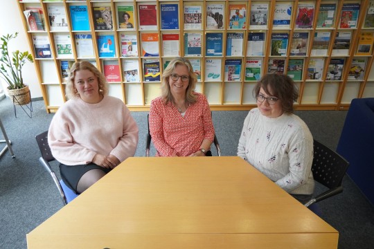 Drei Frauen sitzen an einem Tisch in der Bibliothek. Im Hintergrund ist ein Zeitschriftenregal.